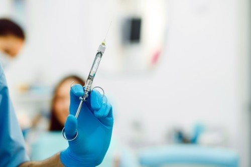 Anaesthesiologist wearing blue medical gloves and holding a syringe in a clinical setting, with a patient and medical equipment blurred in the background